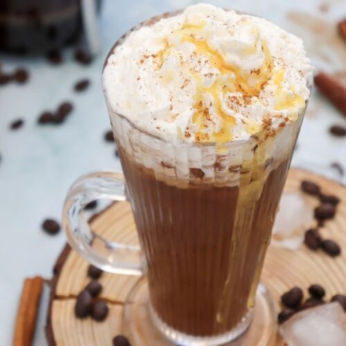 A glass mug of whipped cream-topped coffee sits on a wooden coaster. Drizzled with caramel and sprinkled with cinnamon, the coffee is surrounded by scattered coffee beans and cinnamon sticks on a light surface.
