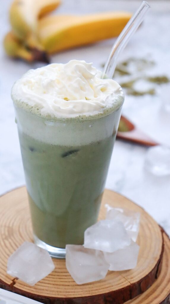A tall glass of iced matcha latte topped with whipped cream and a clear straw. It sits on a round wooden coaster with ice cubes scattered around. Blurred bananas are visible in the background.