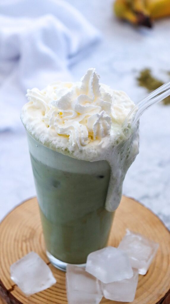 A creamy green beverage topped with generous whipped cream in a glass. Iced cubes are scattered nearby on a wooden coaster. A clear straw is inserted in the drink. A blurred towel and banana are in the background on a marble surface.