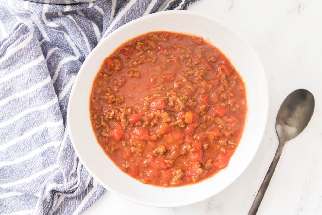 A bowl of chili with ground meat and chunks of tomatoes, placed on a white surface. A gray spoon lies beside it. A blue and white striped cloth is partially visible on the left.