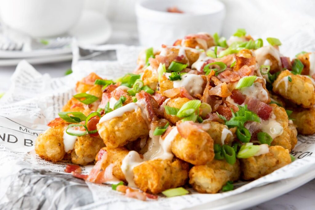 A plate of crispy tater tots topped with melted cheese, crumbled bacon, and chopped green onions. The dish is served on a sheet of newspaper for a rustic presentation. A small white bowl is blurred in the background.