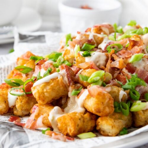 A plate of crispy tater tots topped with melted cheese, crumbled bacon, and chopped green onions. The dish is served on a sheet of newspaper for a rustic presentation. A small white bowl is blurred in the background.