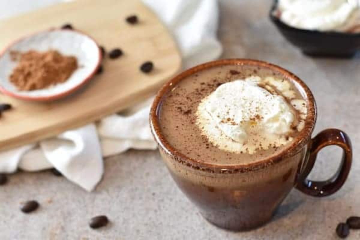A brown mug filled with hot chocolate topped with whipped cream and sprinkled with cocoa powder. In the background, a wooden board with a small bowl of cocoa and scattered coffee beans is visible on a textured surface.