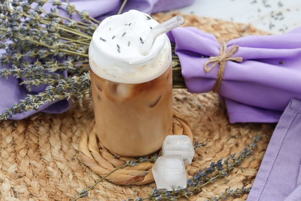 Iced coffee topped with whipped cream and lavender, garnished with lavender sprigs. It sits on a woven mat with ice cubes nearby and is surrounded by purple cloth and lavender bundles.