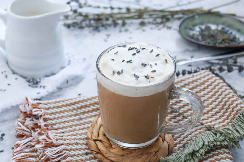 A glass mug of coffee topped with creamy foam and sprigs of lavender on a woven coaster. A striped towel lies underneath, with a white jug and plate of lavender in the background.