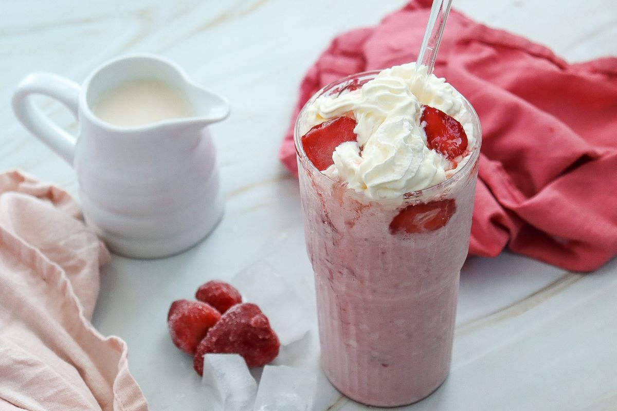 A tall glass of strawberry milkshake topped with whipped cream and sliced strawberries. Beside it, there is a small white pitcher of milk, some frozen strawberries, ice cubes, and a red cloth on a marble surface.