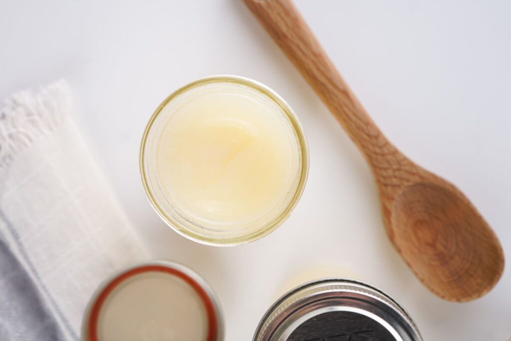 Top view of an open jar of solid beef tallow on a light surface, alongside a wooden spoon and a white cloth. Another jar with a lid is partially visible.