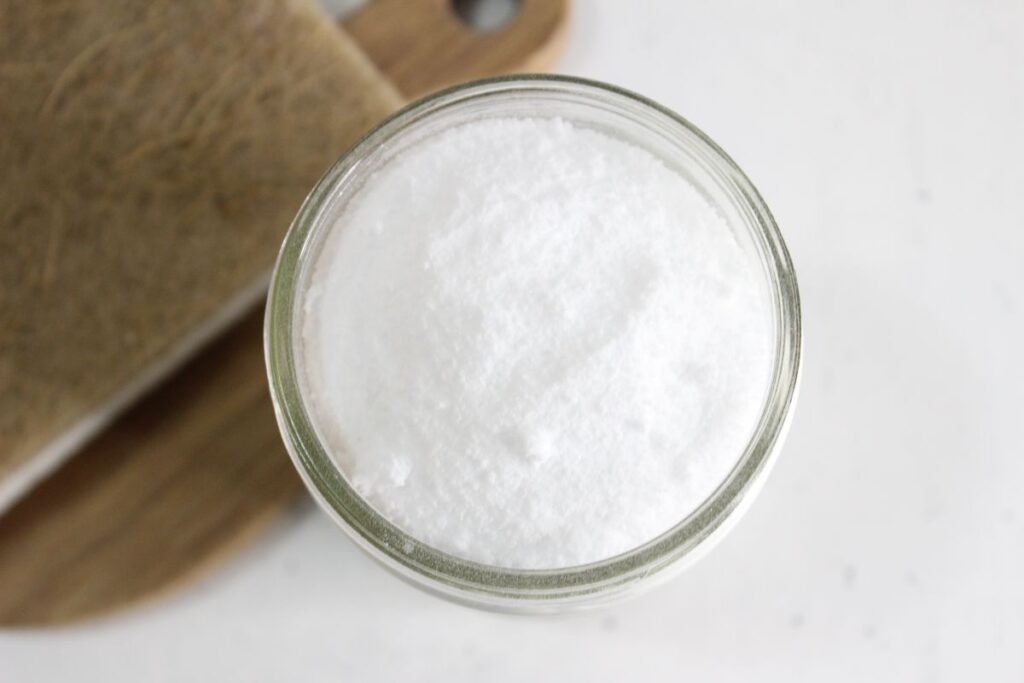 A glass jar filled with white powder, possibly salt, sugar, or flour, is placed on a wooden surface. The jar is viewed from above, showing a smooth, even layer of the powder inside.