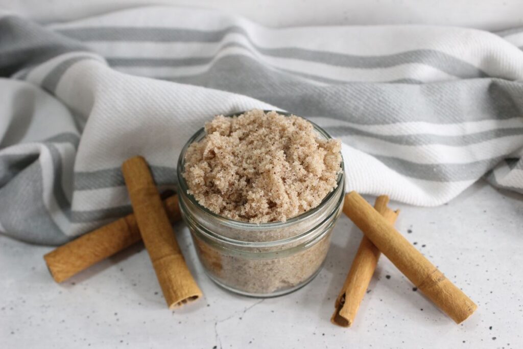A small glass jar filled with a mixture of brown sugar surrounded by three cinnamon sticks. A white and gray striped cloth is in the background, resting on a marble surface.