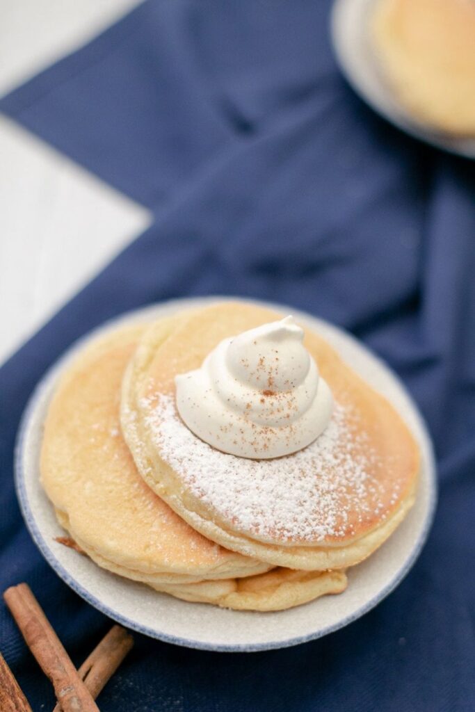 A small stack of pancakes is topped with a dollop of whipped cream and dusted with powdered sugar. The stack is on a gray plate set on a navy blue cloth. Two cinnamon sticks are placed nearby.