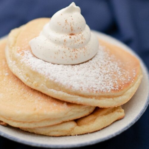 A stack of fluffy pancakes sprinkled with powdered sugar and topped with a swirl of whipped cream, all set on a plate against a dark blue background.