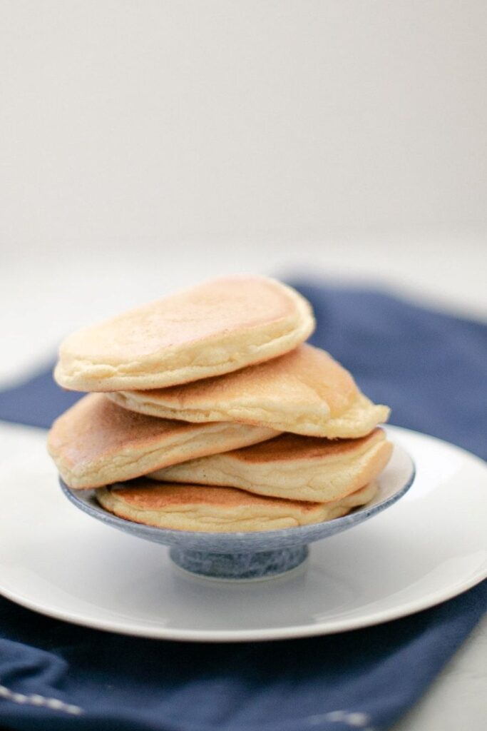 A stack of four fluffy pancakes is placed on a small white and blue plate. The background features a blurred dark blue cloth on a white surface.
