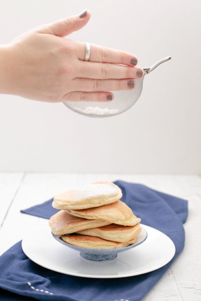 A stack of pancakes on a blue and white plate sits on a dark blue napkin. A hand holds a sieve above the pancakes, sprinkling powdered sugar. The background is light and airy, emphasizing the fluffy pancakes.