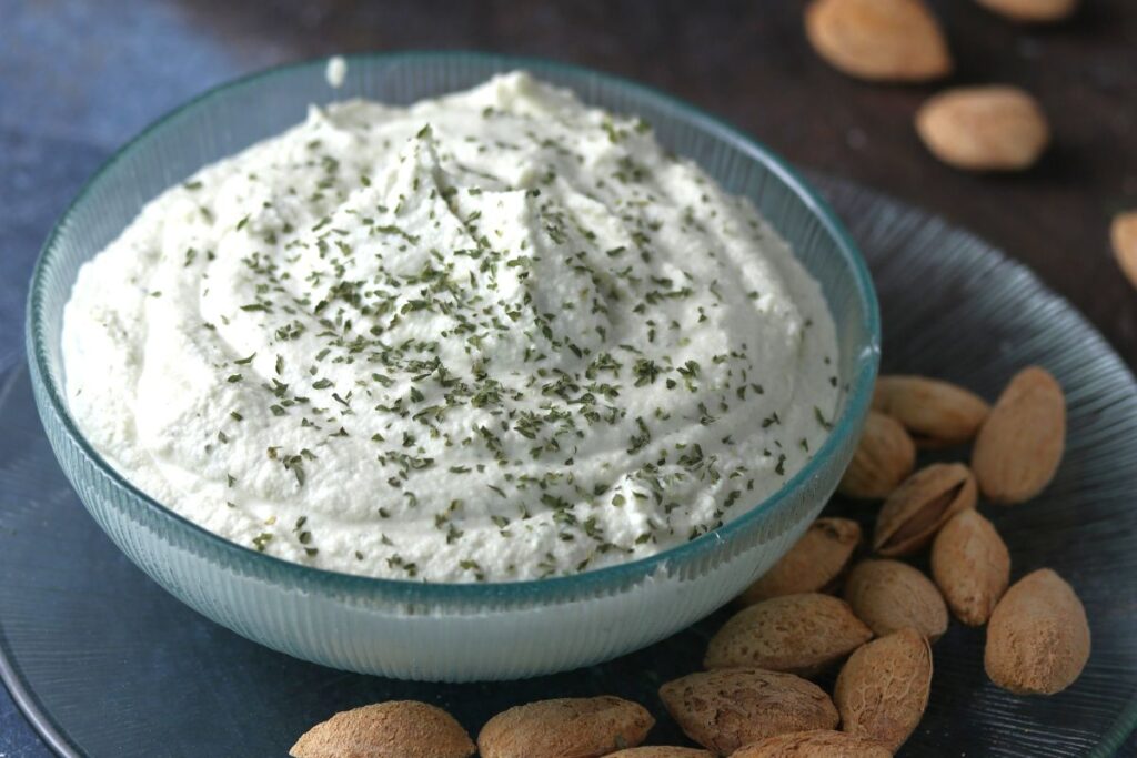 A glass bowl filled with creamy whipped ricotta cheese sprinkled with herbs. The bowl sits on a dark blue plate surrounded by unshelled almonds. The background is a textured dark surface.