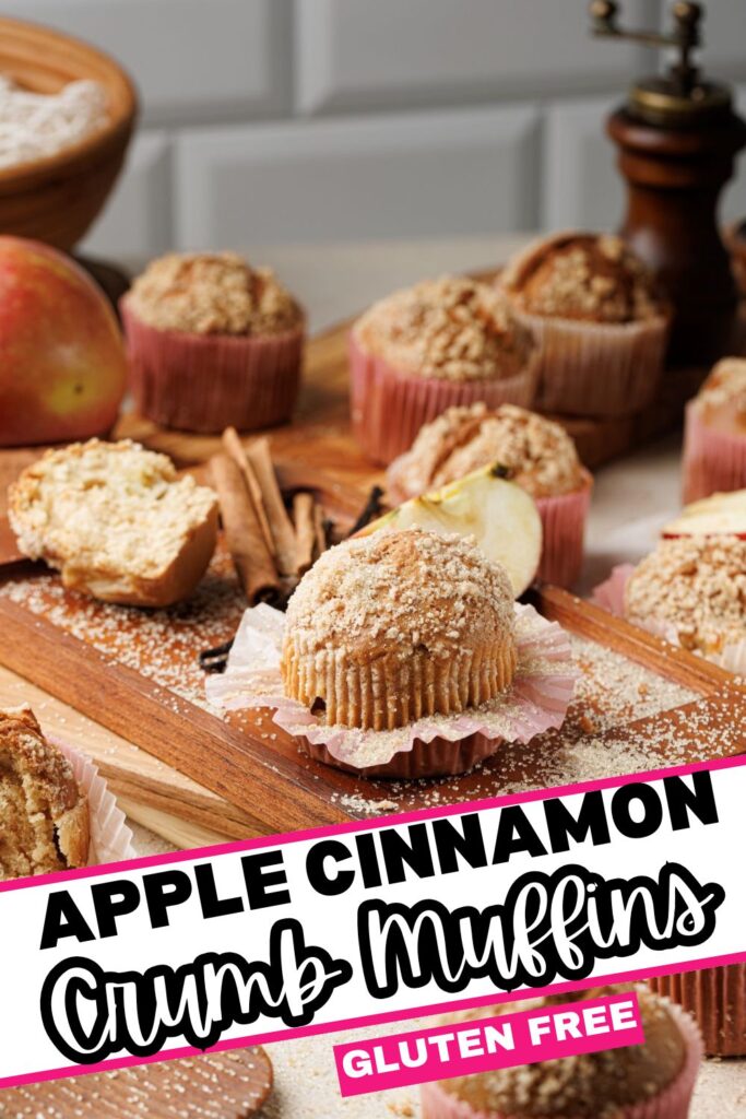 Apple cinnamon crumb muffins displayed on a wooden board, surrounded by apples, cinnamon sticks, and muffin liners. A label at the bottom says "Apple Cinnamon Crumb Muffins - Gluten Free" in bold black and pink text.