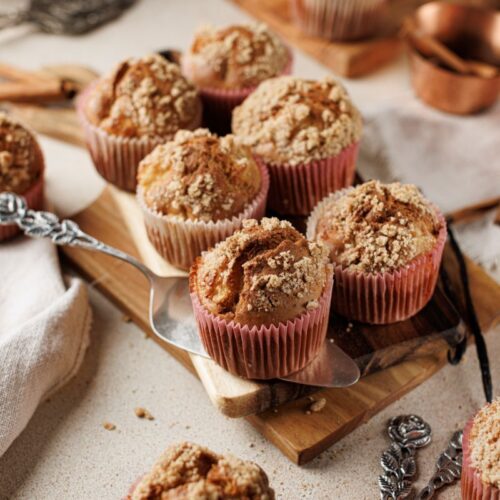 A wooden board topped with apple muffins in pink wrappers, sprinkled with crumb topping. The background includes a whole apple, copper measuring cups, and a white cloth, creating a rustic baking scene.