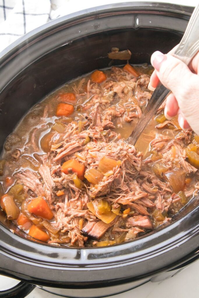 A hand uses a fork to lift shredded beef from a slow cooker filled with meat, carrots, and vegetables in a savory broth. The dish appears hearty and well-cooked.