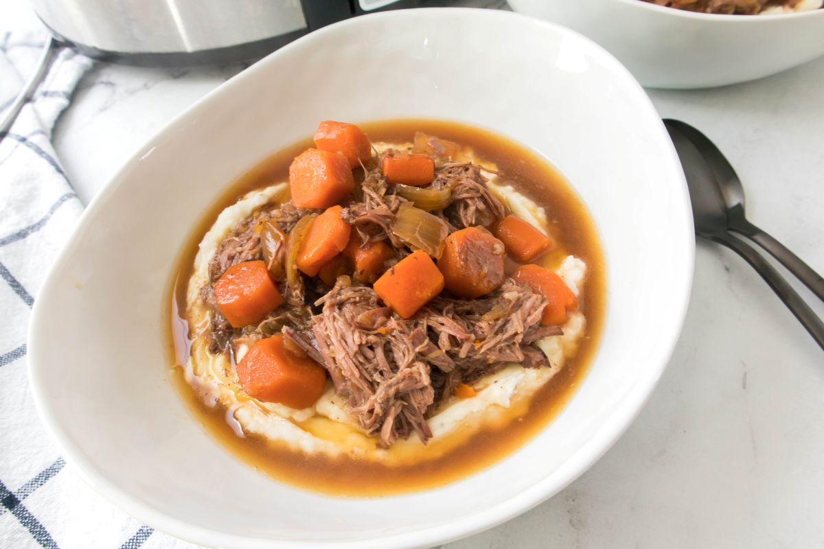 A white bowl filled with shredded beef, carrot chunks, and sauce, served over a bed of mashed potatoes. A black spoon rests beside the bowl, and a checkered cloth and another bowl are in the background.