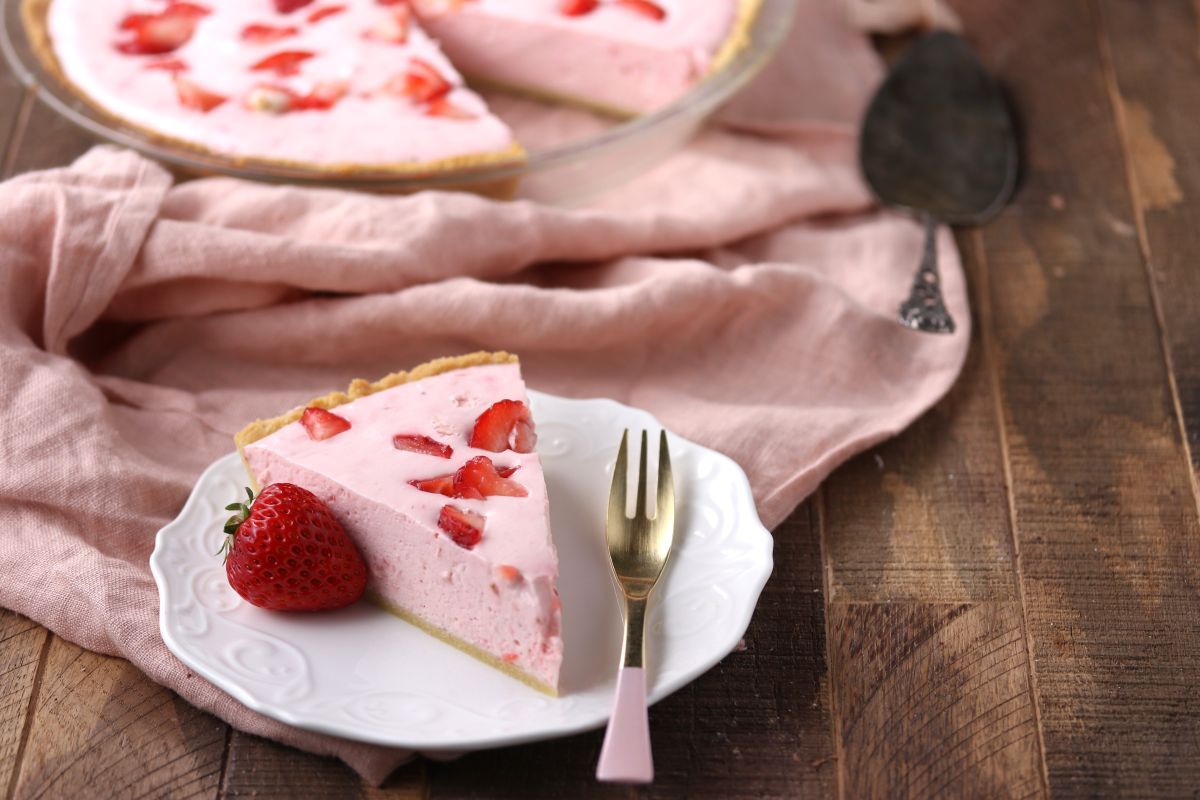 A slice of strawberry cream pie on a white plate with a gold fork, garnished with a fresh strawberry. The pie is pink with strawberry pieces. A whole pie and a vintage serving spoon are in the background, all on a wooden table with a pink cloth.