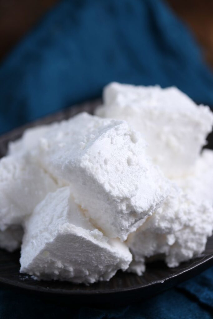 A close-up of a black plate filled with several large, fluffy white marshmallows against a soft, dark blue fabric background. The marshmallows have a powdery texture, appearing soft and light.