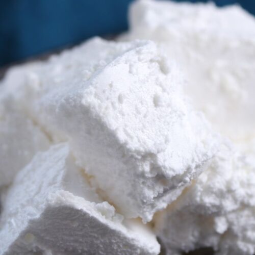 Close-up of several fluffy, white marshmallows piled on a dark plate. The texture appears soft and powdery. A blue cloth is blurred in the background.