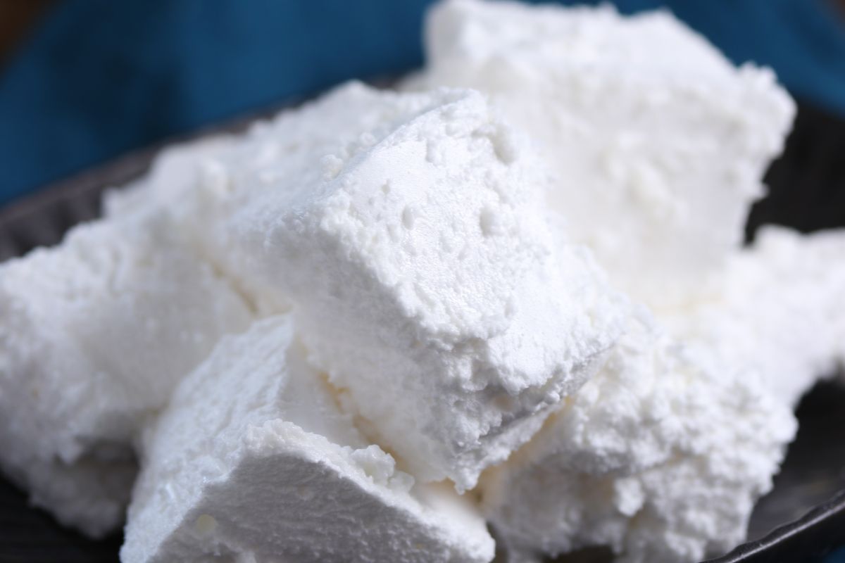 Close-up of several fluffy, white marshmallows piled on a dark plate. The texture appears soft and powdery. A blue cloth is blurred in the background.