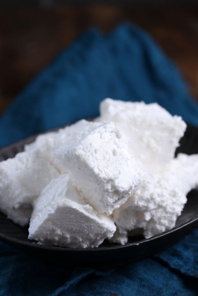 A black bowl filled with several cubes of powdered sugar on a dark blue cloth. The sugar appears soft and fluffy, with a fine texture. The background is out of focus, emphasizing the sugar cubes.