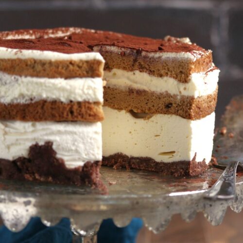 A half-eaten layered tiramisu cake on a cake stand, featuring alternating layers of coffee-soaked sponge, creamy mascarpone, and a dusting of cocoa powder on top. One slice is removed and placed beside it.