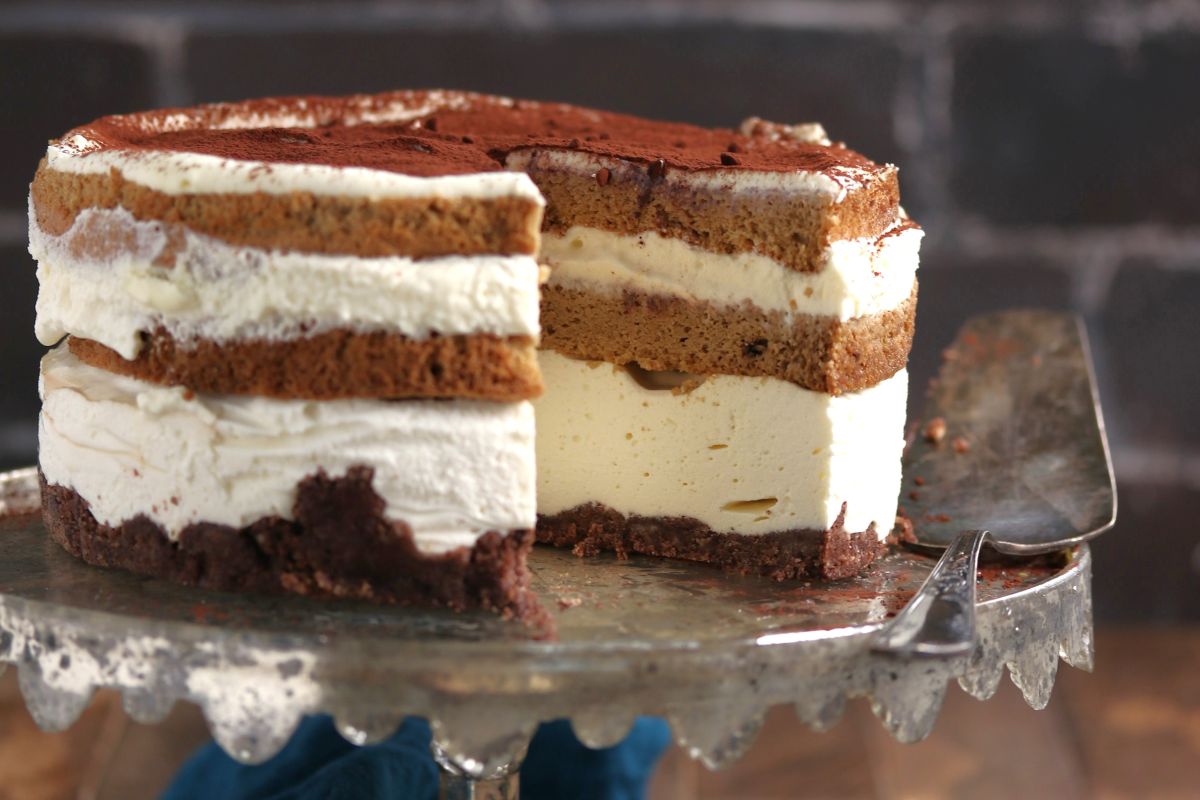 A half-eaten layered tiramisu cake on a cake stand, featuring alternating layers of coffee-soaked sponge, creamy mascarpone, and a dusting of cocoa powder on top. One slice is removed and placed beside it.
