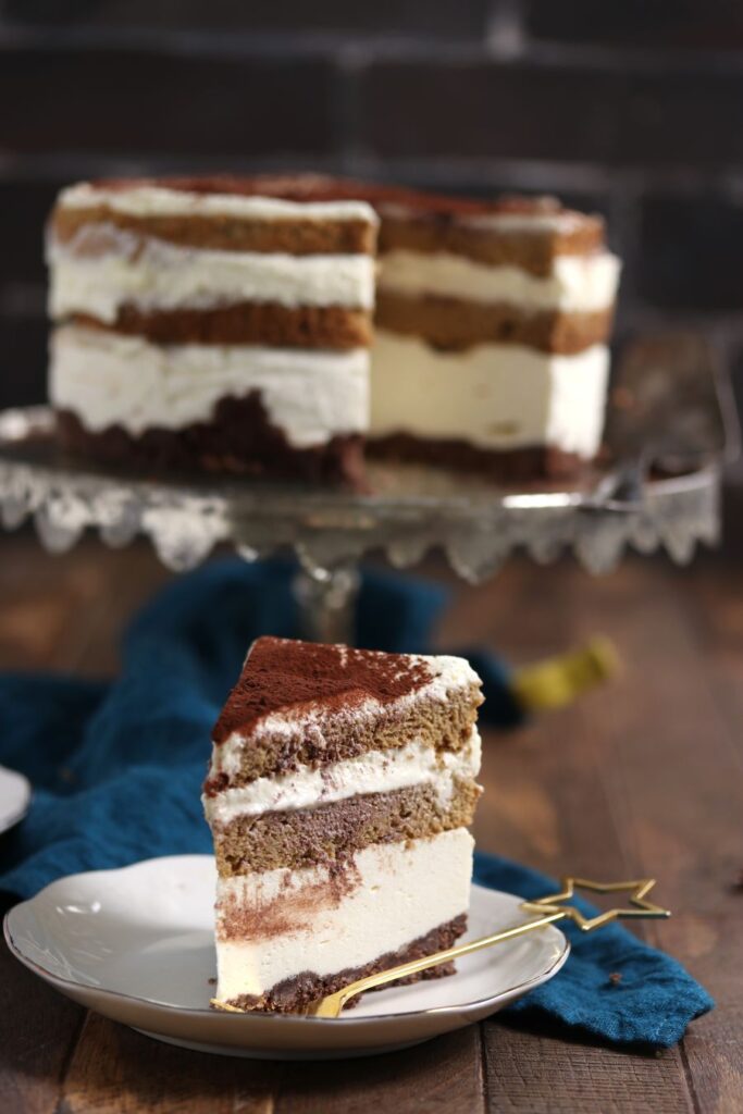 A slice of layered tiramisu sits on a white plate with a gold fork. The dessert features layers of creamy mascarpone, coffee-soaked ladyfingers, and a dusting of cocoa. The full cake is visible in the background on a glass stand.