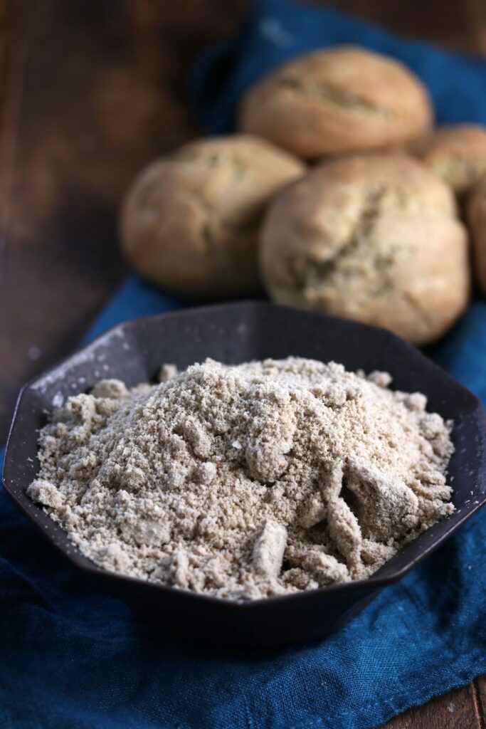 A black bowl filled with buckwheat flour is in the foreground on a blue cloth. In the background, there are four round, rustic bread rolls slightly out of focus. The setting is on a wooden surface.
