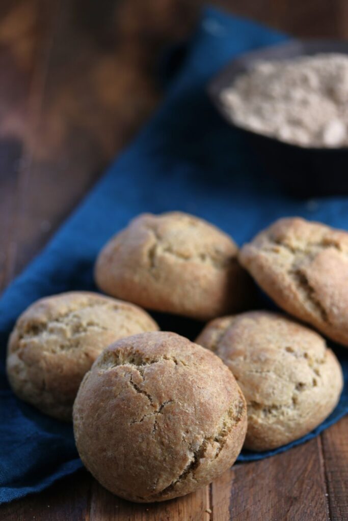 Four round, golden-brown bread rolls rest on a dark blue cloth with a bowl of flour in the background on a wooden surface.