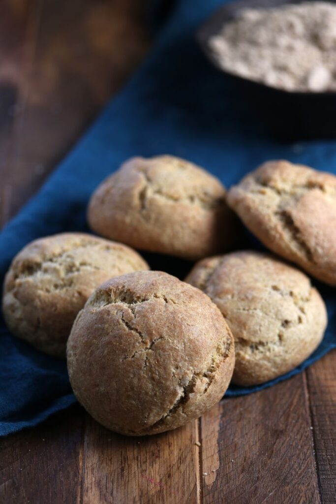 Five round, golden-brown scones on a wooden surface with a blue cloth. A bowl of flour is visible in the background.