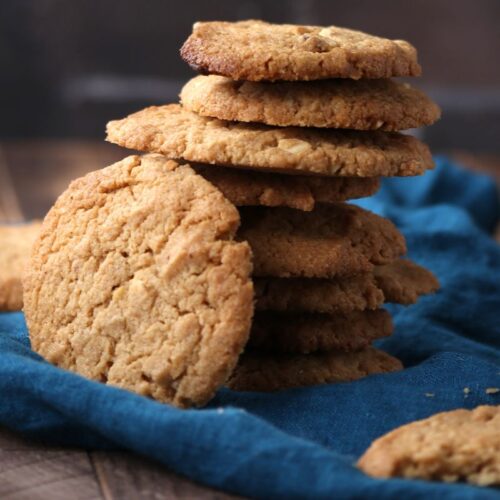 A stack of homemade cookies sits on a blue cloth, with several cookies leaning against the stack and scattered on a wooden surface. The background is blurred and dark.