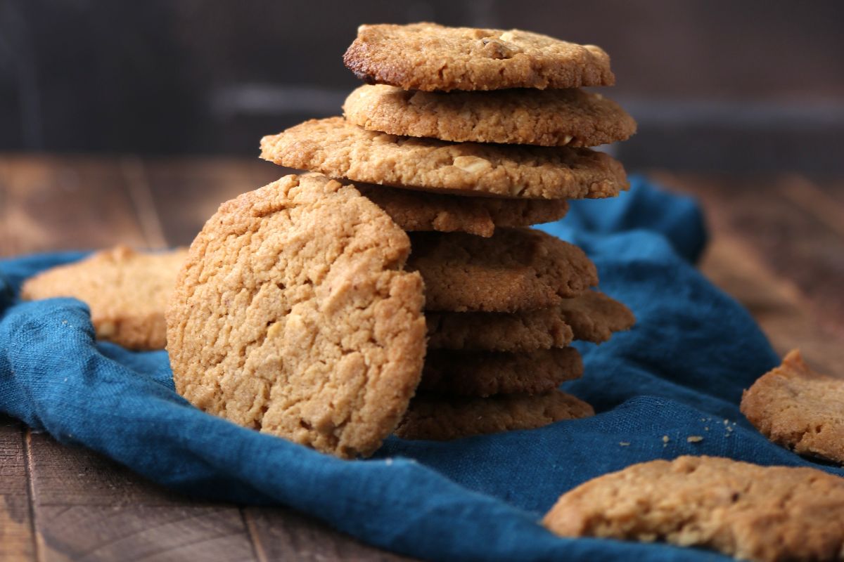 A stack of homemade cookies sits on a blue cloth, with several cookies leaning against the stack and scattered on a wooden surface. The background is blurred and dark.