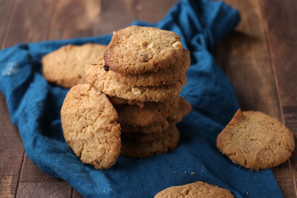 A stack of golden brown cookies sits on a blue cloth atop a rustic wooden surface, with a few cookies scattered around.