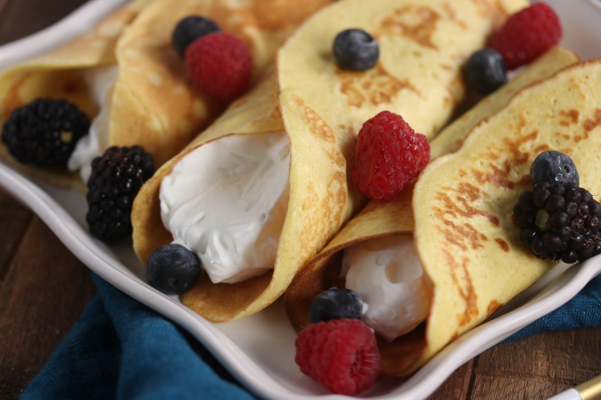 Three rolled crepes filled with whipped cream, garnished with fresh blueberries, raspberries, and blackberries on a white plate. A blue napkin is partially visible under the plate.