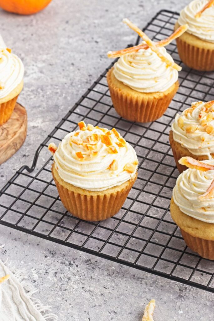 Cupcakes with swirled frosting and orange zest garnish sit on a black cooling rack, with a few cupcakes visible in the background. The surface is light gray and speckled.