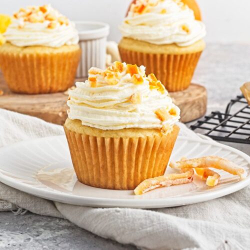 A cupcake with creamy white frosting and orange zest sits on a plate. More cupcakes and orange slices are in the background on a wooden board and cooling rack. The scene is bright and inviting.