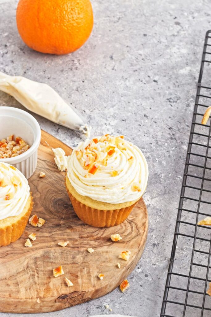 A frosted cupcake topped with orange zest sits on a wooden board with scattered zest, a piping bag, a ramekin of chopped orange, and a cooling rack nearby. An orange is in the background on a gray surface.