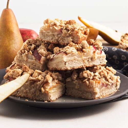 A stack of pear crumble bars on a dark plate, surrounded by fresh pears and slices, with a blue patterned cloth in the background.