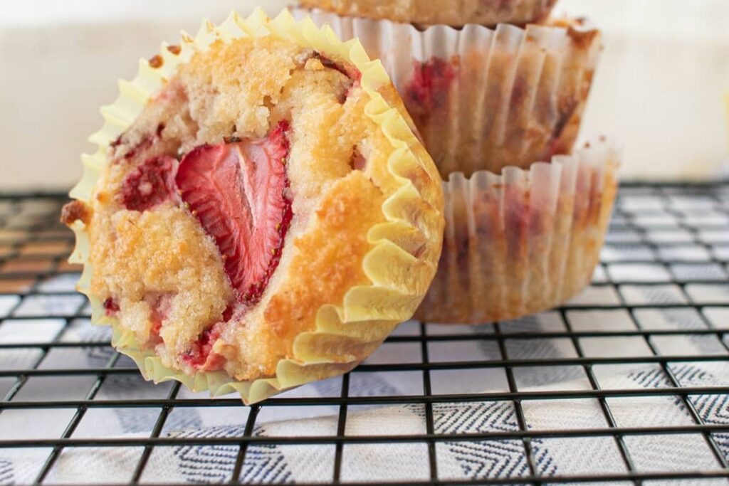 A close-up of strawberry muffins in paper wrappers, one showing a visible strawberry piece on top, resting on a black wire cooling rack with a patterned cloth underneath.