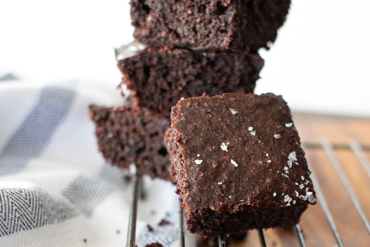 Three thick, fudgy chocolate brownies are stacked on a cooling rack, with one brownie leaning forward. The brownies are topped with flaky sea salt, and a white towel with blue stripes is visible in the background.