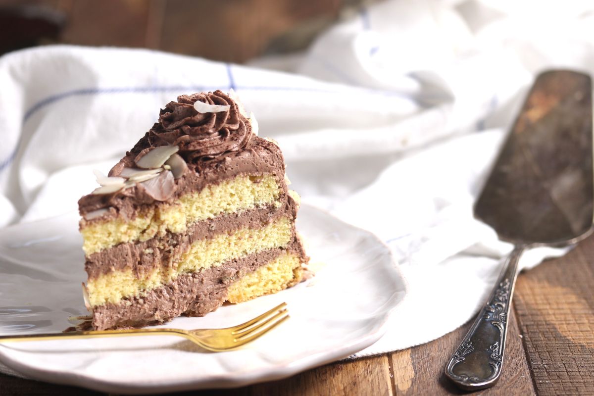 A slice of layered yellow cake with chocolate frosting and almond slices on top sits on a white plate with a gold fork beside it. A cake server and white cloth are in the background on a wooden surface.