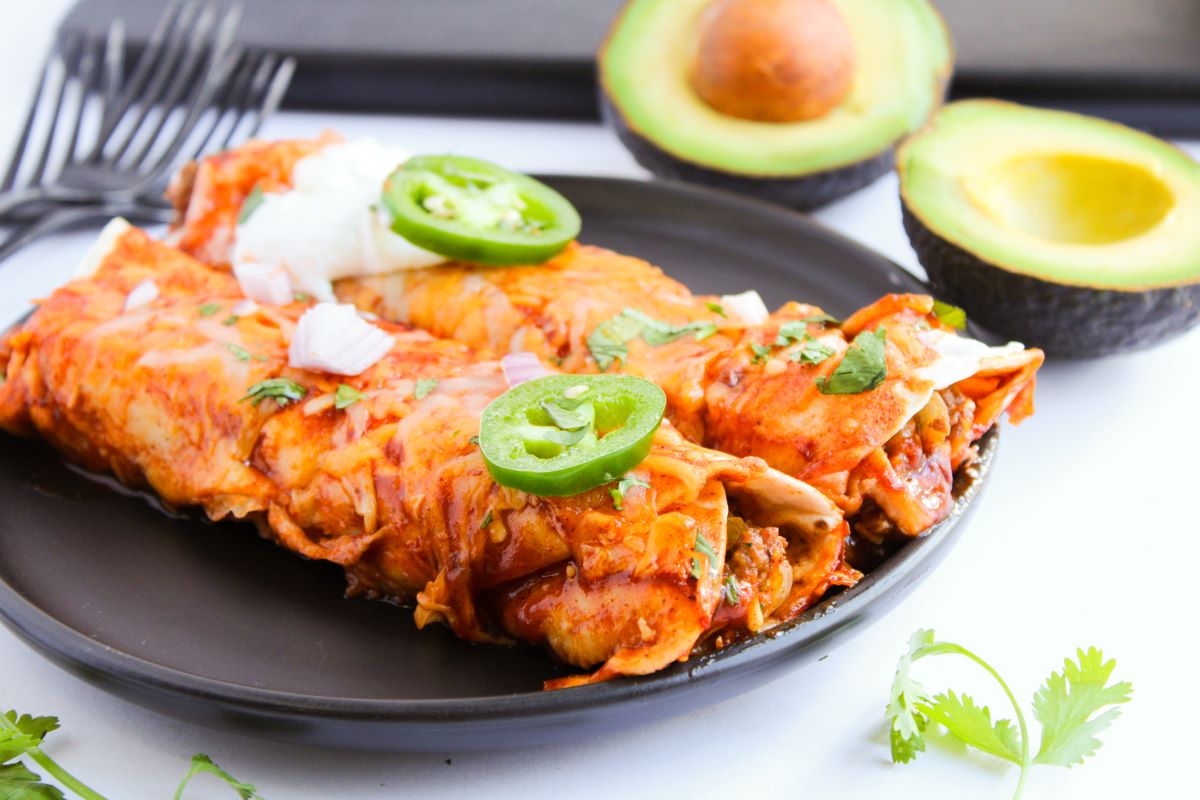 A black plate with two enchiladas topped with sliced jalapeños, sour cream, and onions. In the background, halved avocados and cilantro are visible. Four forks are placed on the side.